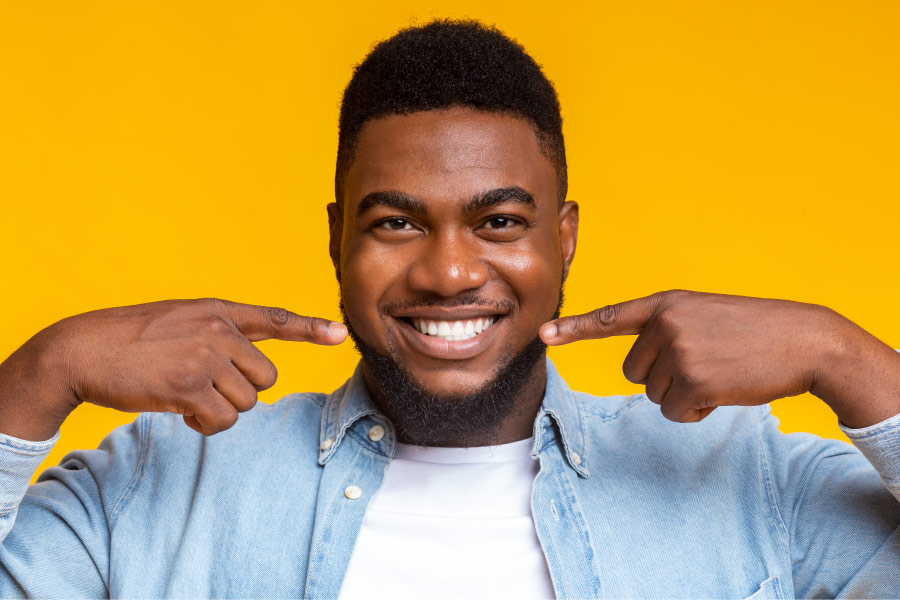 Dark-haired man smiles and points to his healthy teeth and gums against a yellow background in Canton, GA